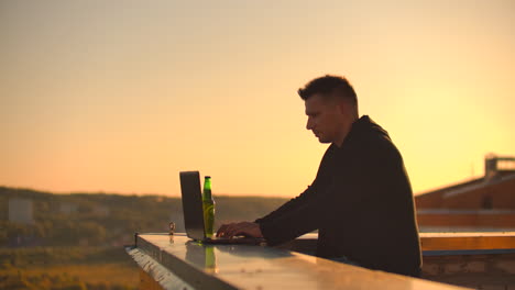 A-male-freelance-programmer-sits-on-a-skyscraper-roof-with-a-laptop-and-beer-typing-code-on-a-keyboard-during-sunset.-Remote-work.